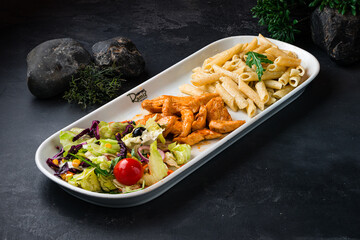 Canvas Print - Penne pasta with fried chicken and a green salad of olives, lettuce, arugula, cherry tomatoes, corn and carrots on a dark table.