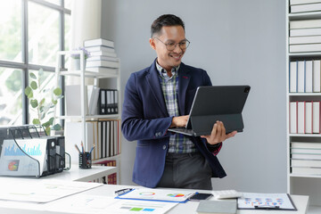 Wall Mural - Asian accountant is standing at his desk in his office, smiling while working on a tablet