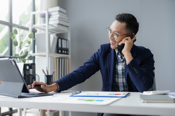 Wall Mural - Asian businessman is working in the office, checking financial data on a digital tablet and talking on his phone