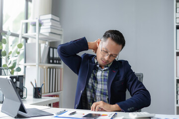 Wall Mural - Young businessman holding his nape and keeping eyes closed, suffering neck pain while sitting at his workplace in the office