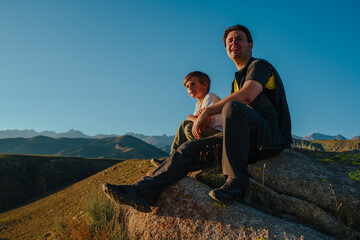 Wall Mural - Father and son sitting on huge stone in the mountains at sunset light