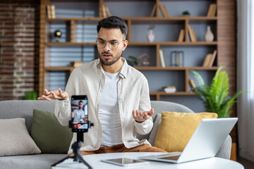 Young man broadcasting live from home using smartphone and laptop for podcasting, discussing important topics in casual setting