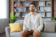 © Tetiana - Young man meditating with closed eyes on a comfortable sofa at home, focusing on relaxation and inner peace