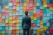 © Vladan - A focused businessman wearing a suit stands in front of a vibrant wall filled with sticky notes, symbolizing brainstorming and creativity in the workplace.