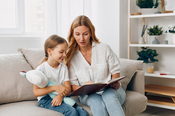 Wall Mural - Mother and daughter bonding on a cozy couch, sharing a storybook and cuddling a stuffed animal