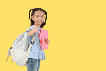 Canvas Print - Cute little happy Asian girl with backpack and notebooks showing thumb-up gesture on yellow background. End of school year