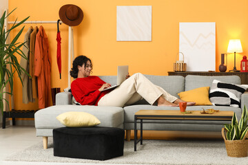 Beautiful young happy African-American woman in autumn outfit with laptop on sofa at home