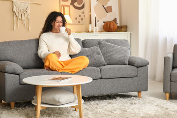 Sticker - Beautiful young happy African-American woman in autumn outfit with cup of tea sitting on sofa at home