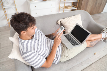 Wall Mural - Handsome young man with air conditioner remote control and laptop lying on sofa at home