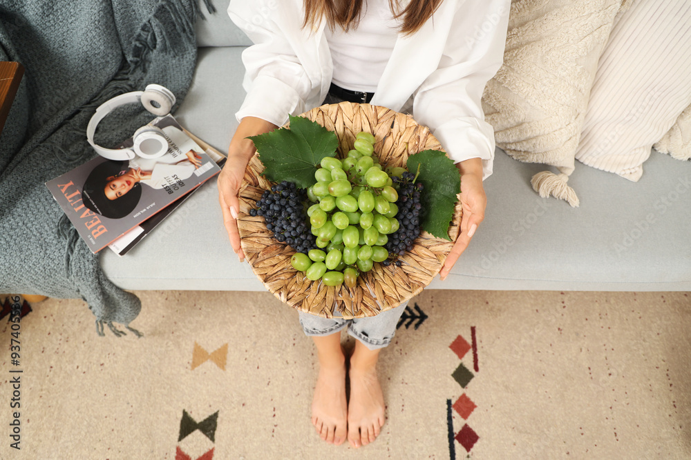 Young woman with grapes sitting on sofa at home, top view