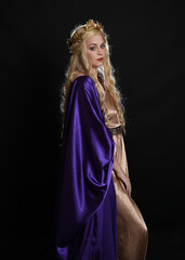 Wall Mural - Close up portrait of beautiful blonde female model wearing purple and gold silken toga robes with royal crown, historical fantasy goddess costume. Isolated on dark studio background.