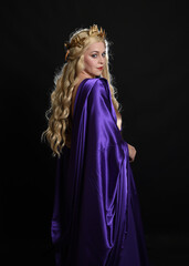 Canvas Print - Close up portrait of beautiful blonde female model wearing purple and gold silken toga robes with royal crown, historical fantasy goddess costume. Isolated on dark studio background.