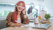 © wattana - Smiling young woman sitting at a desk with a professional microphone, writing note and preparing for the podcast episode.