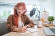 © wattana - Smiling young woman sitting at a desk with a professional microphone, writing note and preparing for the podcast episode.