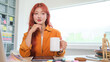 © wattana - Confident young stylish woman with vibrant red hair holding a coffee mug sitting at her creative workspace.