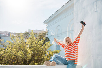 Wall Mural - A woman is sitting on a ledge with her phone in her hand. She is wearing a striped shirt and blue jeans. The scene is set in a city with a tree in the background.