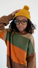 A young girl with curly hair showcases her fashionable side, wearing an oversized brown and green sweater paired with a yellow beanie, exuding confidence and charm