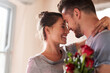 © Chanelle2000/peopleimages.com - Flowers, happy couple and hug in home for love, connection and bonding together with present. Affection, man and woman with red roses for relationship, commitment or anniversary celebration in house