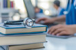 © Amazing Kingdom - Close-up of stethoscope resting on a stack of medical books with a doctor using a computer in the background. Medical knowledge and learning concept.