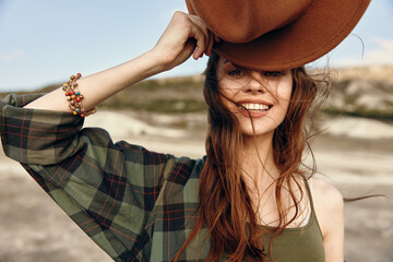 Wall Mural - Stylish woman in plaid shirt and hat poses with hands on head in desert setting