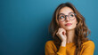 © khonkangrua - Thoughtful young woman wearing glasses, looking upwards, posing against a blue background, reflecting a contemplative and curious mood.