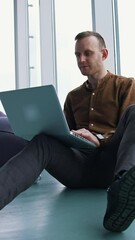 Wall Mural - Young freelancer working on a laptop. Man in shirt and trousers sitting on a floor and using a gadget to work in office. Panoramic windows view. Vertical video