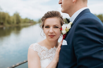 A bride and groom are posing for a picture by a body of water. The bride is wearing a white dress and the groom is wearing a blue suit. Scene is romantic and happy