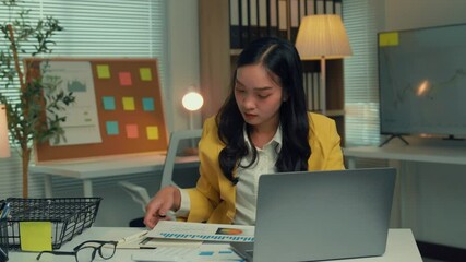 Poster - Young businesswoman is reviewing documents at her desk in her office at night. She is working on her laptop and analyzing paperwork