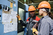© pressmaster - Engineer and Factory Worker discussing printed plans in an industrial setting, both wearing protective helmets and gloves. Engaging in a technical discussion with detailed diagrams pinned to a board