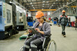 © pressmaster - Worker in wheelchair using tablet for managing processes in modern industrial facility. Colleague in background walking between machines, both wearing safety gear