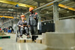 © pressmaster - Two workers in protective gear collaborating in factory environment, one person seated in wheelchair holding clipboard and device, surrounding machinery and metal components visible
