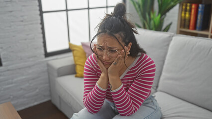 Poster - Concerned young hispanic woman sitting on a white couch in an apartment, looking distressed