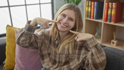 Canvas Print - A smiling caucasian woman sits in a cozy living room pointing to her cheeks in a playful pose.