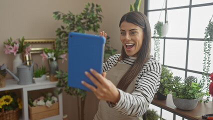 A happy young hispanic woman takes a selfie indoors surrounded by houseplants and gardening tools.