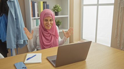 Wall Mural - A smiling woman in a pink hijab engaging in a video call on a laptop at a home office setting