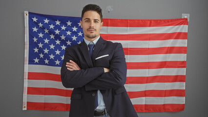 A confident young hispanic man in a suit standing with crossed arms in an office with an american flag in the background.