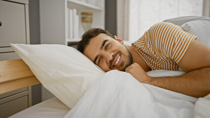 Wall Mural - A handsome young hispanic man with a beard smiles while lying in bed in a cozy bedroom.