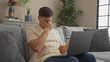© Krakenimages.com - Young, hispanic man sitting indoors on a sofa, rubbing his eyes while using a laptop, in a living room of a house.