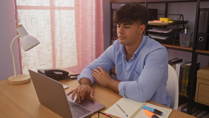 Wall Mural - A handsome young hispanic man in an office setting, working on a laptop while surrounded by office supplies and furniture in a well-lit room.