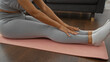 © Krakenimages.com - African american woman seated on a pink yoga mat at home stretching while wearing a gray workout outfit.