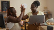 © Krakenimages.com - Two black women bakers high-fiving in a bakery shop, one holding a laptop and the other a coffee cup, displaying joyful and collaborative team spirit in an indoor setting