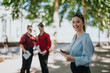 © qunica.com - Young businesswoman holding documents outdoors while colleagues are interacting in the background under the shade of trees on a sunny day.