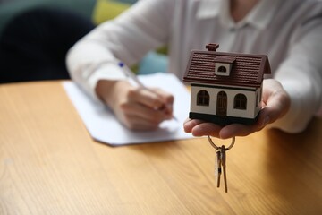 Wall Mural - Woman working with document while holding house model and key at wooden table indoors, closeup