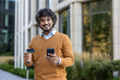 © Liubomir - Smiling man holding credit card and phone outside modern building, suggesting concepts of online payment, business travel, and digital communication. Casual attire and outdoor setting