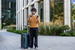 © Liubomir - Businessman standing outside modern office with suitcase, holding credit card and phone, focused on work. Urban setting with sunlight reflecting on glass windows, creating professional atmosphere.