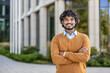 © Liubomir - Confident young man with curly hair smiling outside modern office building. Wearing orange sweater with arms crossed, expressing positivity and professionalism. Reflects successful business outlook