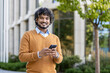 © Liubomir - Cheerful young man holding phone outdoors, dressed in casual autumn clothing. Standing confidently near modern office environment with greenery, embodying relaxed professionalism and comfort.