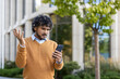 © Liubomir - Young man feeling frustrated while looking at phone outside modern office building. He gestures with confusion, expressing annoyance. Wearing brown sweater, standing in urban environment under tree.