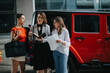 © qunica.com - Three businesswomen stand by a red car, engaged in a discussion, reviewing documents and notes.