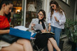 © qunica.com - Three businesswomen holding a meeting outdoors, focusing on reviewing and discussing documents. Collaboration and teamwork in a professional setting.